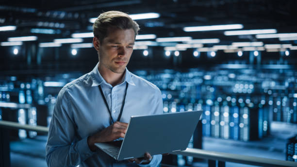 istock Handsome Smiling IT Specialist Using Laptop Computer in Data Center. Succesful Businessman and e-Business Entrepreneur Overlooking Server Farm Cloud Computing Facility. 1336250793