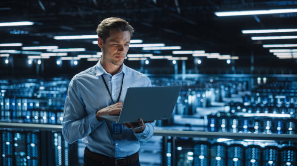 Handsome Smiling IT Specialist Using Laptop Computer in Data Center. Succesful Businessman and e-Business Entrepreneur Overlooking Server Farm Cloud Computing Facility. Handsome Smiling IT Specialist Using Laptop Computer in Data Center. Succesful Businessman and e-Business Entrepreneur Overlooking Server Farm Cloud Computing Facility. data center stock pictures, royalty-free photos & images