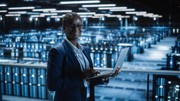 centre de données: programmeuse utilisant un ordinateur portable, souriant et regardant la caméra. dans cloud computing server farm system administrator travaillant sur la maintenance informatique pour iaas, saas, paas. - data center computer programmer women photos et images de collection