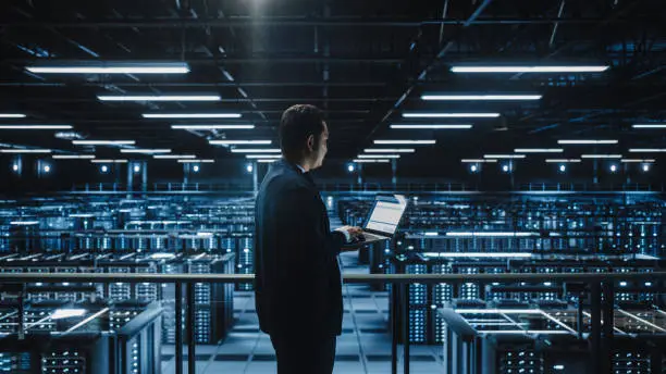 Photo of Data Center Male Administrator Using Laptop Computer. Maintenance Specialis working in Cloud Computing Facility on Cyber Security and Network Protection. Server Farm Analytics. Medium Wide