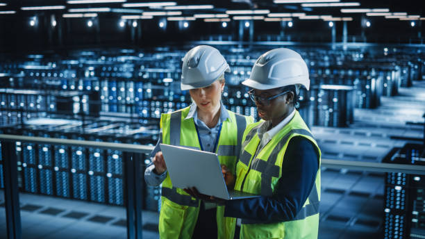 istock Data Center IT Specialist and System administrator Talk, Use Laptop Computer, Wearing Safety West. Server Cloud Farm Facility with Two Female Information Technology Engineers Doing Maintenance. 1336250844