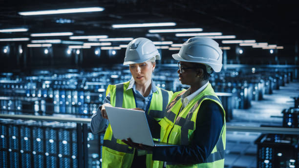 istock Data Center IT Specialist and System administrator Talk, Use Laptop Computer, Wearing Safety West. Server Cloud Farm Facility with Two Female Information Technology Engineers Doing Maintenance. 1336250845