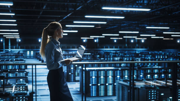 istock Data Center Female IT Specialist Uses Laptop Computer. Information Technology Engineer Overseeing and Walking Through Cloud Computing Server Farm Maintenance and Monitoring. 1336250950