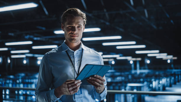 retrato de un apuesto y sonriente especialista en ti que usa una tableta en el centro de datos, mirando la cámara. exitoso ingeniero de negocios electrónicos masculino que trabaja en big server farm cloud computing facility. - engineer fotografías e imágenes de stock