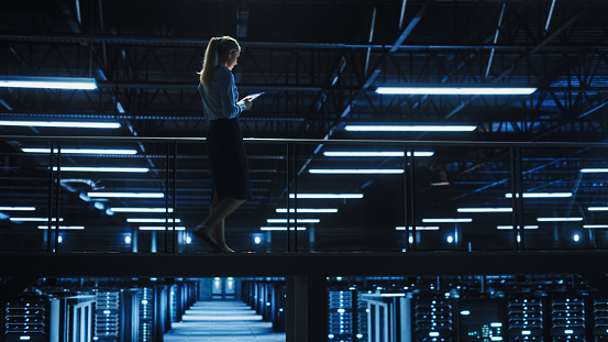 Female IT Specialist Using Tablet Computer in Data Center, Walking on a Bridge Overlooking Gigantic Server Farm Cloud Computing Facility. Succesful Businesswoman, e-Business Entrepreneur.