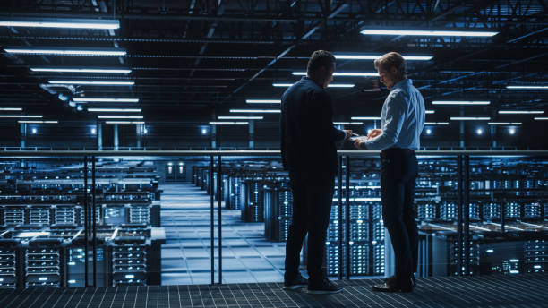 istock Data Center IT Specialist and e-Business Enrepreneur Talk, Use Laptop. Two Information Technology Professionals Walking on Bridge Overlooking Cloud Computing Server Farm. 1336250990
