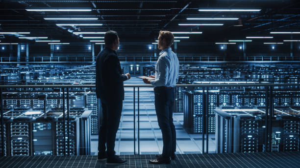 istock Data Center IT Specialist and e-Business Enrepreneur Talk, Use Laptop. Two Information Technology Professionals Walking on Bridge Overlooking Cloud Computing Server Farm. 1336250997