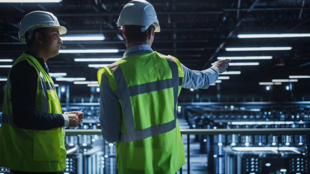 istock Data Center IT Specialist and System administrator Talk, Use Tablet Computer, Wearing Safety Wests. Server Clod Farm with Two Information Technology Engineers checking Cyber Security. 1336251007