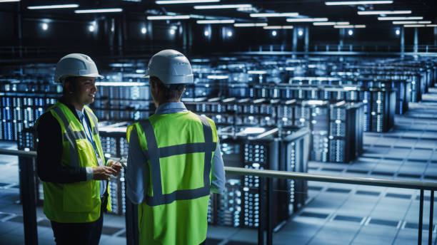 istock Data Center IT Specialist and System administrator Talk, Use Tablet Computer, Wearing Safety Wests. Server Clod Farm with Two Information Technology Engineers checking Cyber Security. 1336251018