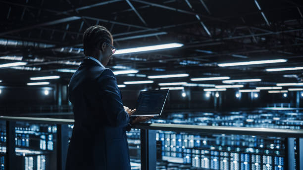 istock Female IT Specialist Using Laptop Computer in Data Center, Walking on a Bridge Overlooking Big Server Farm Cloud Computing Facility. Businesswoman, e-Business Entrepreneur. 1336251019