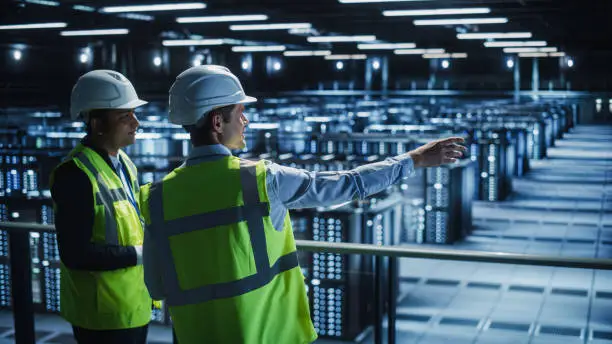 Photo of Data Center IT Specialist and System administrator Talk, Use Tablet Computer, Wearing Safety Wests. Server Clod Farm with Two Information Technology Engineers checking Cyber Security.