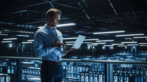 istock Handsome Smiling IT Specialist Using Laptop Computer in Data Center. Succesful Businessman and e-Business Entrepreneur Overlooking Server Farm Cloud Computing Facility. 1336251027