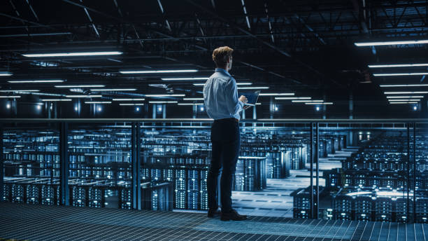 istock Handsome Smiling IT Specialist Using Tablet Computer in Data Center. Succesful Businessman and e-Business Entrepreneur Overlooking Big Server Farm for Cloud Computing. 1336251029