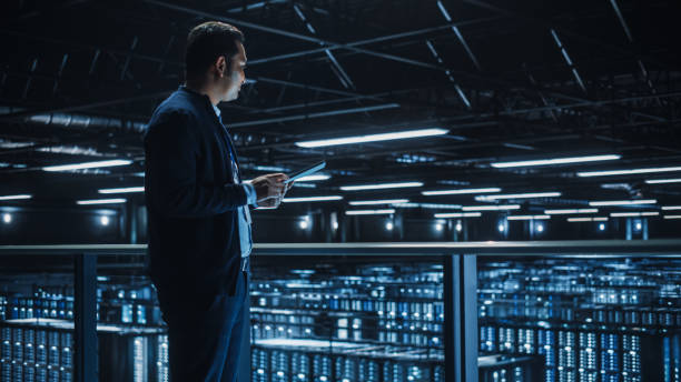 istock Handsome Smiling IT Specialist Using Tablet Computer in Data Center. Succesful Businessman and e-Business Entrepreneur Overlooking Server Farm Cloud Computing Facility. 1336251032