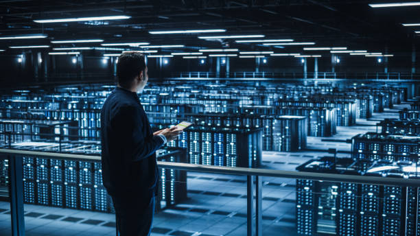 istock Handsome Smiling IT Specialist Using Tablet Computer in Data Center. Succesful Businessman and e-Business Entrepreneur Overlooking Server Farm Cloud Computing Facility. 1336251041
