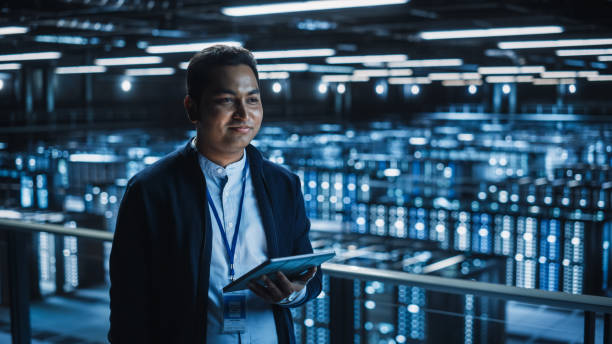 istock Handsome Smiling IT Specialist Using Tablet Computer in Data Center. Succesful Businessman and e-Business Entrepreneur Overlooking Server Farm Cloud Computing Facility. 1336251045