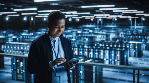 istock Handsome Smiling IT Specialist Using Tablet Computer in Data Center. Succesful Businessman and e-Business Entrepreneur Overlooking Server Farm Cloud Computing Facility. 1336251053