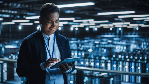 przystojny uśmiechnięty specjalista it korzystający z tabletu w data center. udany biznesmen i przedsiębiorca e-biznesowy overlooking server farm cloud computing facility. - smart tablet zdjęcia i obrazy z banku zdjęć