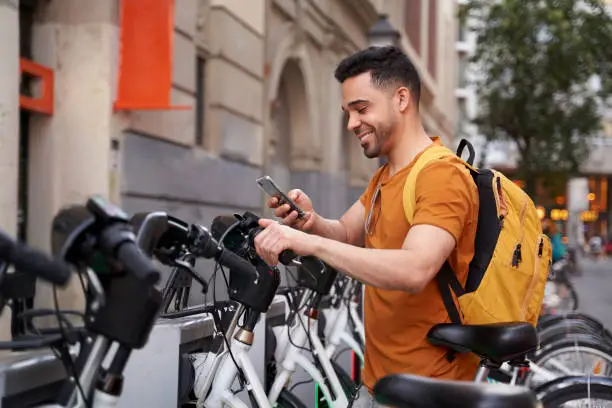 Positive young Latino man stands by the parking lot with a rental bike and uses a smartphone with a smile on his face. Eco-friendly transport concept. High quality photo