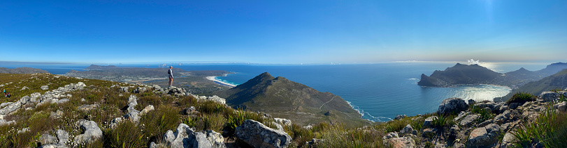 A fit woman enjoys hiking in nature, she is standing on a rock overlooking the bay of water below her. Panoramic of a Woman standing on a rock and looking over False Bay and Cape Point