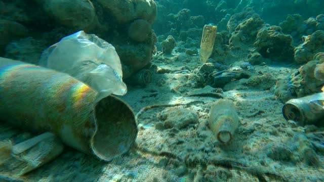 Beautiful coral reef covered with plastic and other garbage, colorful tropical fish swims over this debris. Plastic pollution of the Ocean. Camera slowly moving forward above seabed with plastic trash