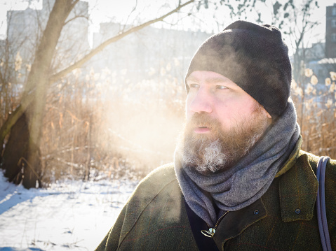 Man breathing in winter forest. Adult man wearing knit hat, breathing cold air. Bearded man relaxing on winter walk in snowy forest, candid capture, lifestyle scene