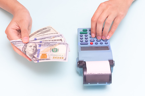 Payment for purchases in cash, dollars. Close-up of a hand giving cash and hand typing the amount, counting at the cash register against a blue background. Business concept, retail, online sale.