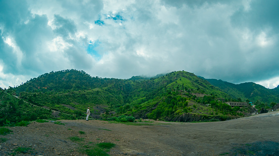 Wide-angle view from Solan, Himachal Pradesh in the rainy season. The sky is full of clouds, the mountains are green with trees, and a man stands alone at a distance looks tiny in the huge landscape.