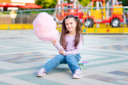 a child girl in an amusement park in the summer eats cotton candy on a skateboard and smiles with happiness near the carousels, the concept of summer holidays and school holidays