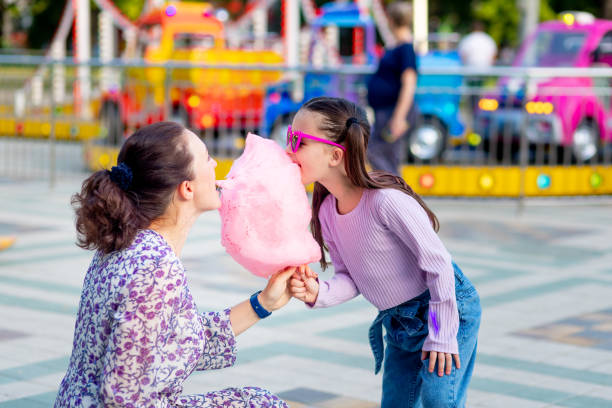 a child girl with her mother in an amusement park in the summer eating cotton candy and ice cream near the carousels, fooling around and laughing, the concept of family weekends and school holidays a child girl with her mother in an amusement park in the summer eating cotton candy and ice cream near the carousels, fooling around and laughing, the concept of family weekends and school holidays child cotton candy stock pictures, royalty-free photos & images