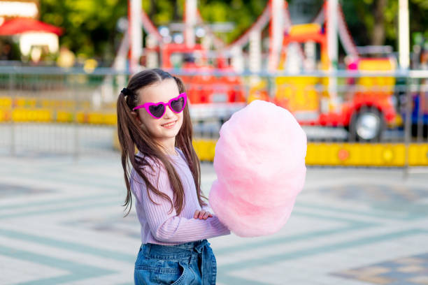 a child girl in an amusement park in the summer eats cotton candy near the carousels in sunglasses, the concept of summer holidays and school holidays a child girl in an amusement park in the summer eats cotton candy near the carousels in sunglasses, the concept of summer holidays and school holidays candyfloss stock pictures, royalty-free photos & images