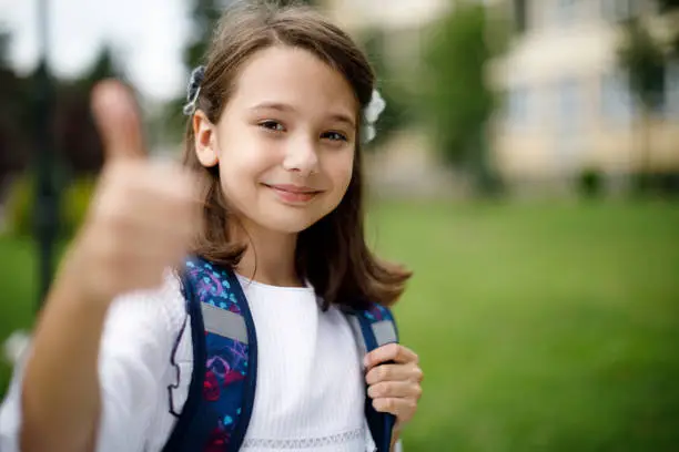 Cute smiling happy schoolgirl gesturing thumb up in front of the school