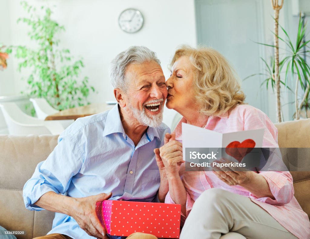 gift woman man couple happy love happiness present kiss romantic smiling together box wife husband elderly old senior mature retired Smiling senior woman receives present box and kissing her husband for giving her a present at home Valentine's Day - Holiday Stock Photo