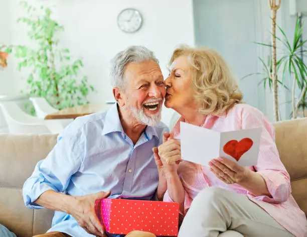 Smiling senior woman receives present box and kissing her husband for giving her a present at home