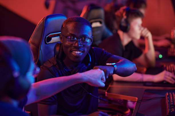 uśmiechnięty młody czarnoskóry mężczyzna w okularach siedzący na krześle komputerowym i uderzając pięścią członka zespołu, gdy grają razem w grę wideo - friendship video game young adult party zdjęcia i obrazy z banku zdjęć