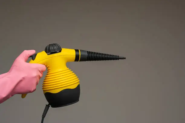 Male hand in rubber glove holding a small steam pressure cleaner. Close up studio shot, isolated on gray background.
