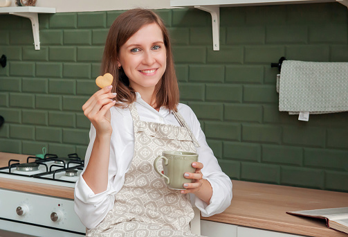 Happy slender young woman in an apron with a mug and cookies is sitting at home in the kitchen. Young hostess in the kitchen. Cooking cookies at home. Pastry chef girl.