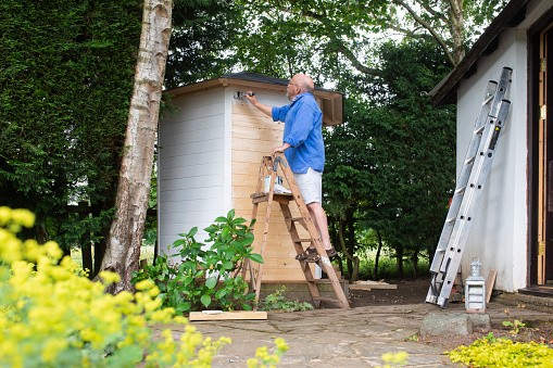 A shot of a senior man on step ladders painting his newly constructed summer house in his garden.