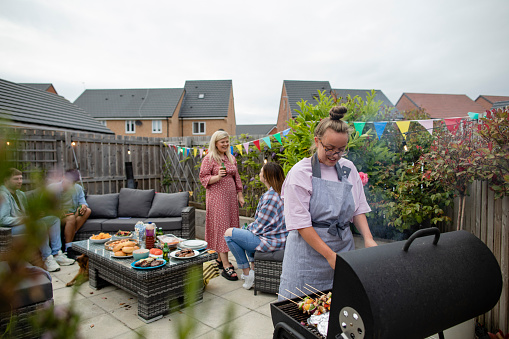 Group of friends having a social gathering outdoors in the North East of England. They are sitting together sharing food on an outdoor patio while the host cooks food on a bbq.
