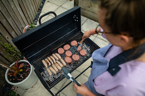 High angle view of a woman in her garden in the North East of England cooking on a BBQ, flipping burgers. She is throwing a social gathering in summer.