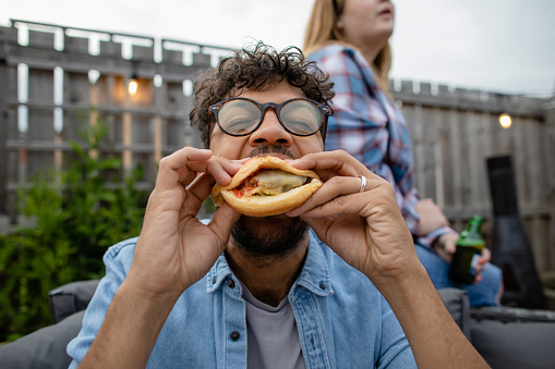 Close up of a man eating a burger while at a bbq social gathering outdoors in the North East of England. Friends are sitting together sharing food on an outdoor patio.