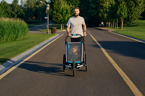 Young father with his kid in a jogging stroller during jogging in a public park. Togetherness dad with son during active running, happy fatherhood