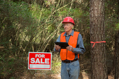 Construction manager marking trees for be cut.  He carries a digital tablet and wears a safety vest and hard hat.  For sale sign is on the premises.