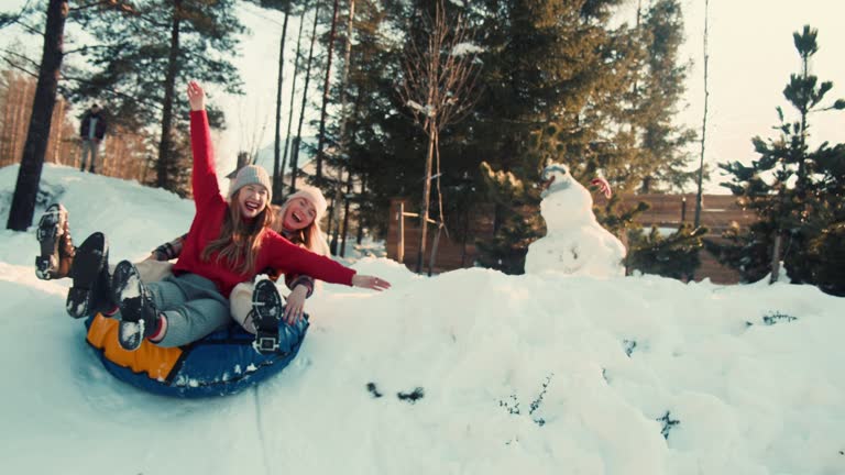 Winter holiday fun. Two beautiful excited happy women smile sledging on snow forest slope towards camera slow motion.