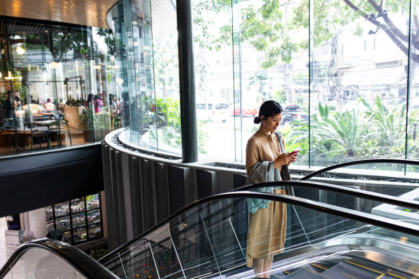 Asian Woman With a Smartphone Riding on an Escalator Asian woman using her mobile phone on a shopping mall escalator thailand mall stock pictures, royalty-free photos & images