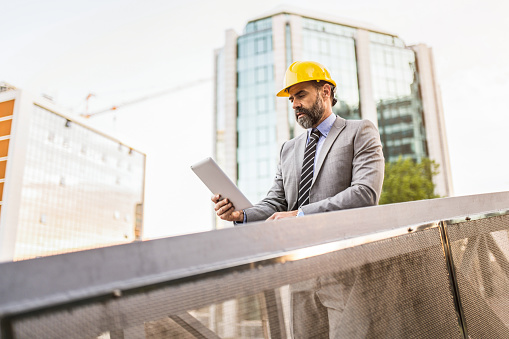 Mature engineer using a digital tablet on a bridge at construction site and examining a new building project with work helmet on.