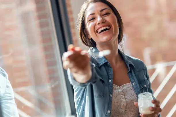 Portrait of beautiful smiling woman having fun while feeding yogurt to camera at home.