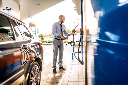 Full length photo of mature businessman filling up car tank with fuel at gas station.