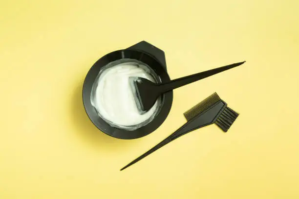 Photo of set for home or salon hair dyeing in the hands of a woman with gloves. Brushes and bowl for hair dye on yellow background