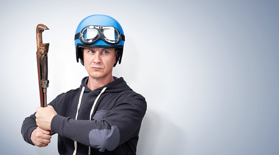 Confident man in sweatshirt and motorcycle helmet with goggles holding a large adjustable rusty wrench, on blue background.